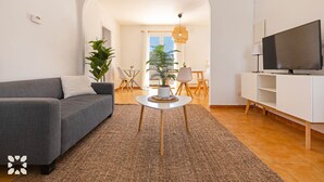 Ett modernt vardagsrum med en grå soffa, en tv på ett vitt tv-bord, ett litet runt bord med en växt och en matplats i bakgrunden.