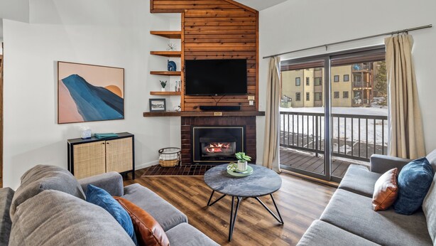 A modern living room with a fireplace, a large TV, and a balcony with a view of buildings. - Renovated 3-Bedroom Condo at Timber Run – Spacious Mountain Retreat with Prime L (Steamboat Springs)