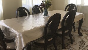 A dining area with a table covered by a plastic tablecloth, surrounded by dark wooden chairs. A vase with flowers is placed on the table. - Beautiful, tranquil Home. 8 minutes from the International Airport (Vieux Fort)