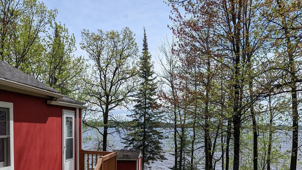 Una baita rossa con veranda in legno e un piccolo deposito, circondata da alberi e un lago.