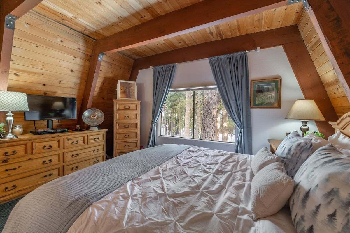 A cozy bedroom with a wooden ceiling, a bed with a patterned duvet, a dresser with drawers, a television, and a window with curtains.