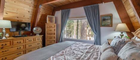 A cozy bedroom with a wooden ceiling, a bed with a patterned duvet, a dresser with drawers, a television, and a window with curtains.