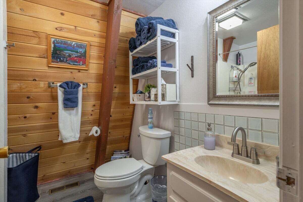 A wooden-paneled bathroom with a toilet, a shelf with towels, a sink, and a mirror.