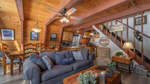 A cozy cabin interior with a wooden ceiling, a sofa, a dining table, and a kitchen area.