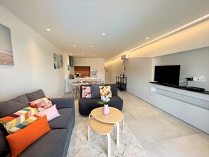 Ein modernes Wohnzimmer mit einem grauen Sofa, bunten Kissen, einem kleinen Holztisch und einem an der Wand befestigten Fernseher.