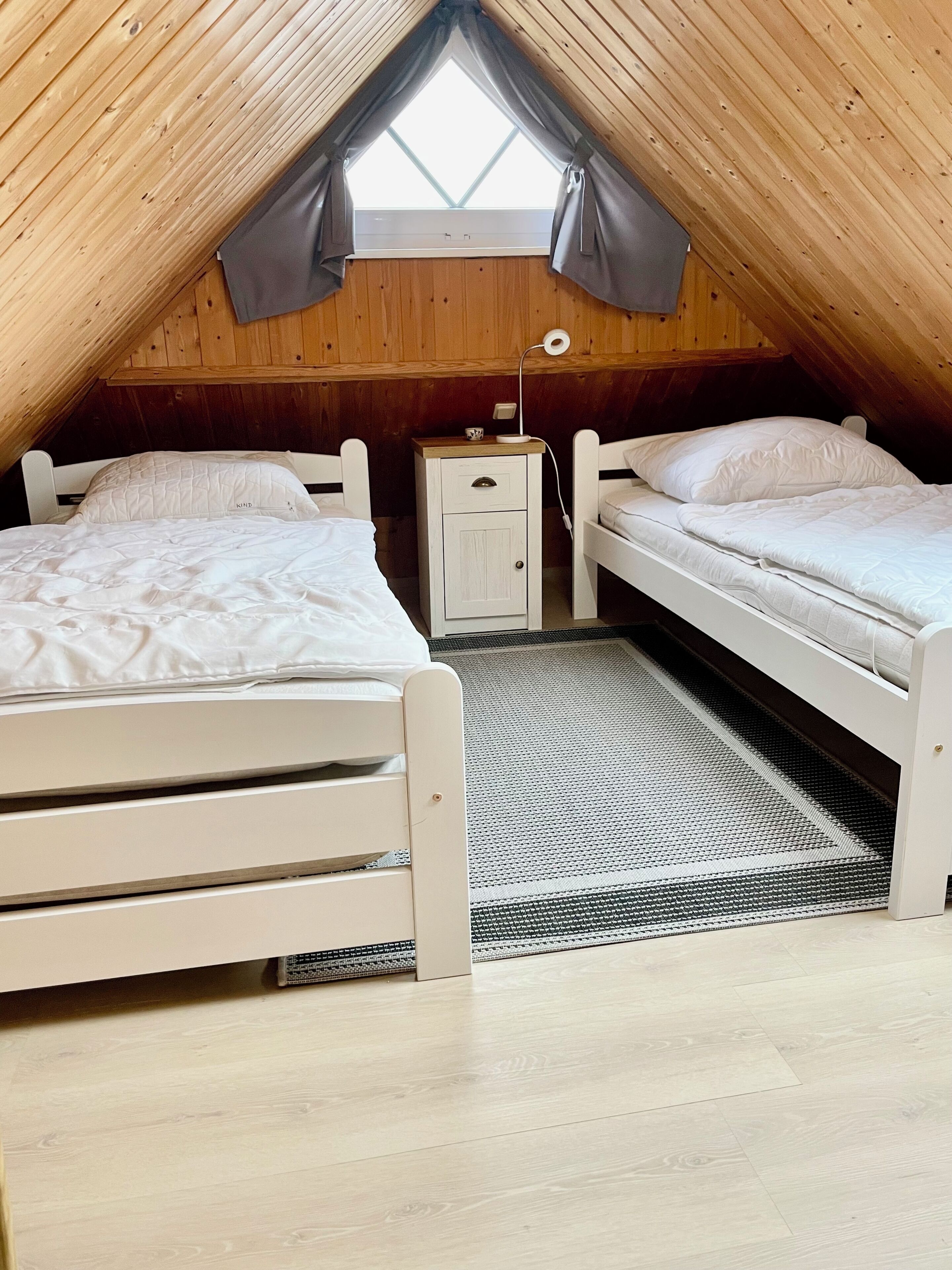 Een kleine slaapkamer op zolder met twee bedden, een nachtkastje, een raam met gordijnen en een houten plafond.