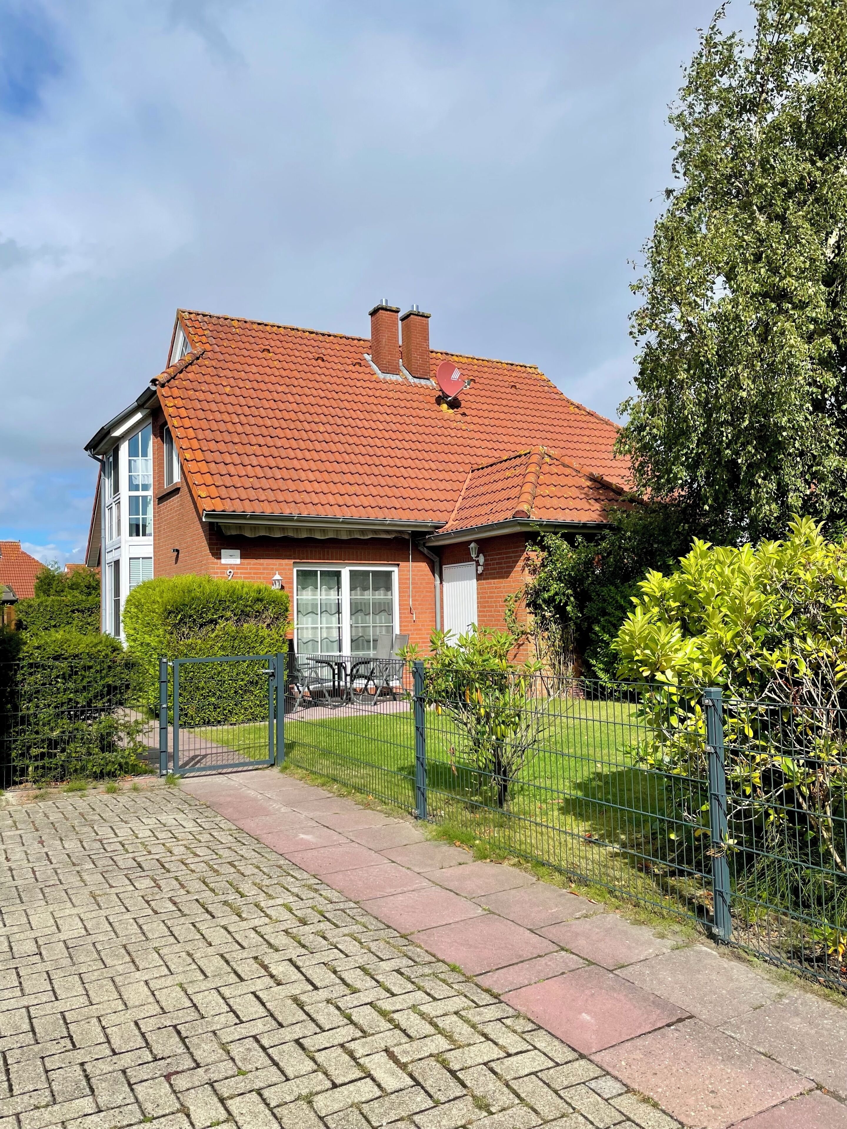 Une maison à deux étages, avec un toit recouvert de tuiles rouges, une façade en briques et une allée d’accès fermée par un portail.