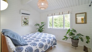 Un dormitorio con cama, dos plantas en macetas, ventana con cortinas, lámpara colgante y aire acondicionado instalado en la pared.