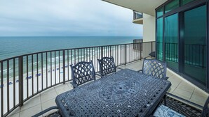 A balcony with a view of the beach and ocean, a table with chairs, and a glass door.