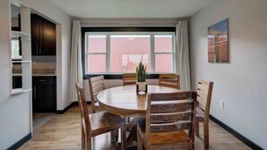 A dining area with a round wooden table and chairs, a potted plant, and a view of a pink building through large windows. - Ranch Guest House with Mountain Views (Belgrade)