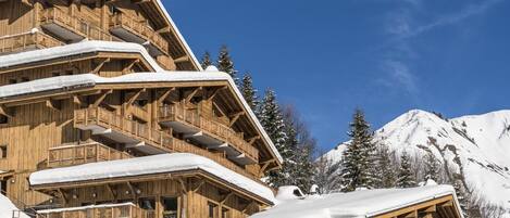Un chalet en bois à plusieurs étages, doté de balcons, qui se dresse sur fond de montagnes enneigées.