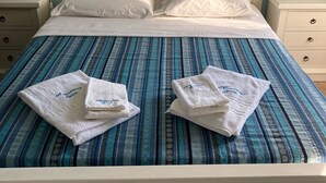 Un llit ben arrenjat amb un llençol blaus i blancs a ratlles, dues tovelloles blanques amb text brodat i una paret amb elements decoratius, incloent-hi un objecte amb forma de cor.