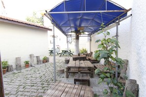 Zona de pati exterior amb taules i bancs de fusta, una teulada blava i plantes en testos.