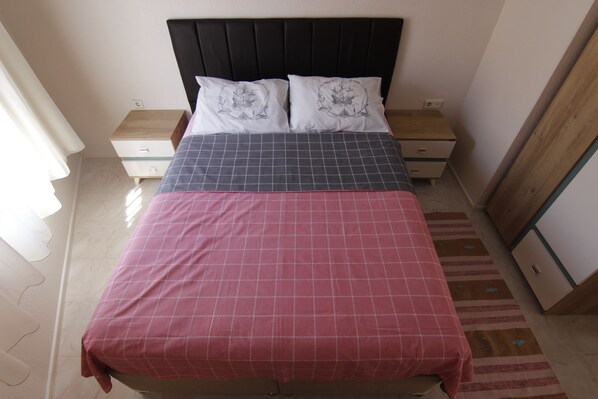 A bed with a red and grey checkered bedspread, two white pillows with a ship design, and a black headboard. There are wooden nightstands with drawers and a wooden wardrobe to the right. - Antik Ev Apart (Cesme)