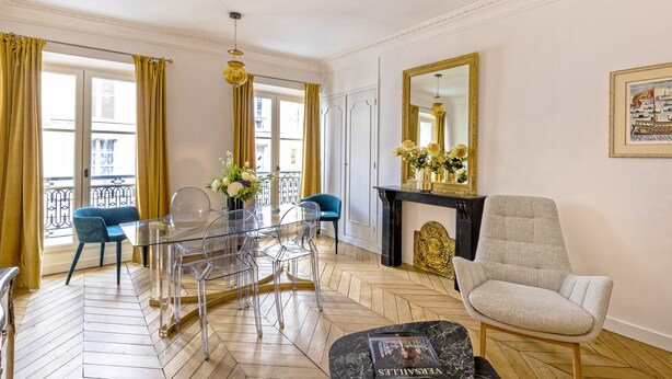 A spacious room with a herringbone floor, a glass dining table with chairs, a blue armchair, a grey armchair, and a fireplace with a mirror above it.