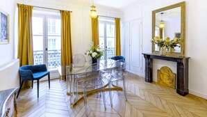 A room with a glass table surrounded by chairs, a fireplace with a mirror, and a view of the outside through large windows with yellow curtains.