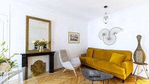 A living room with a herringbone floor, a yellow sofa, a white armchair, a black coffee table, a fireplace, and a large mirror.