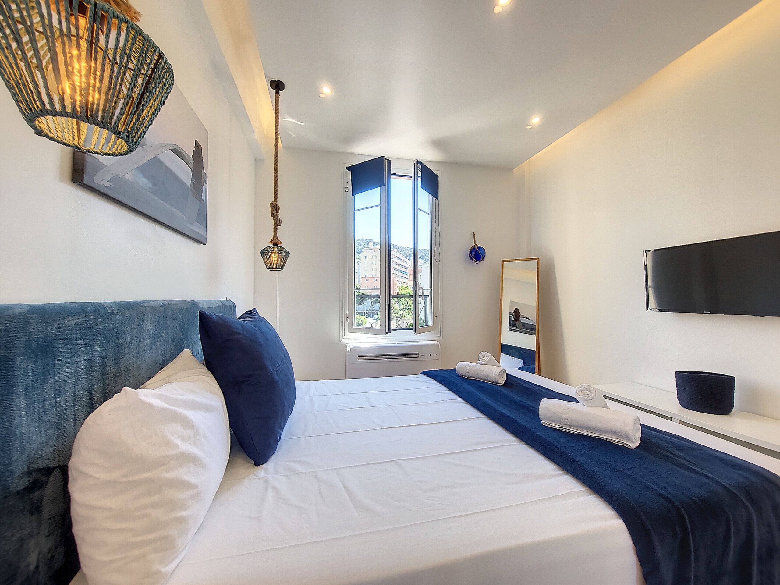 A modern hotel room with a large bed, a flat-screen TV, a window with a view, and a ceiling light fixture.