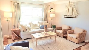 Una sala de estar acogedora con un sofá, dos sillones, una mesita de centro y una mesita auxiliar. Un modelo de velero cuelga en la pared, y se observa un reloj también en la pared.