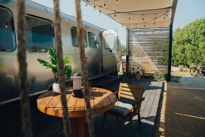 A camper van parked on a deck with a round wooden table, chairs, and a lantern.