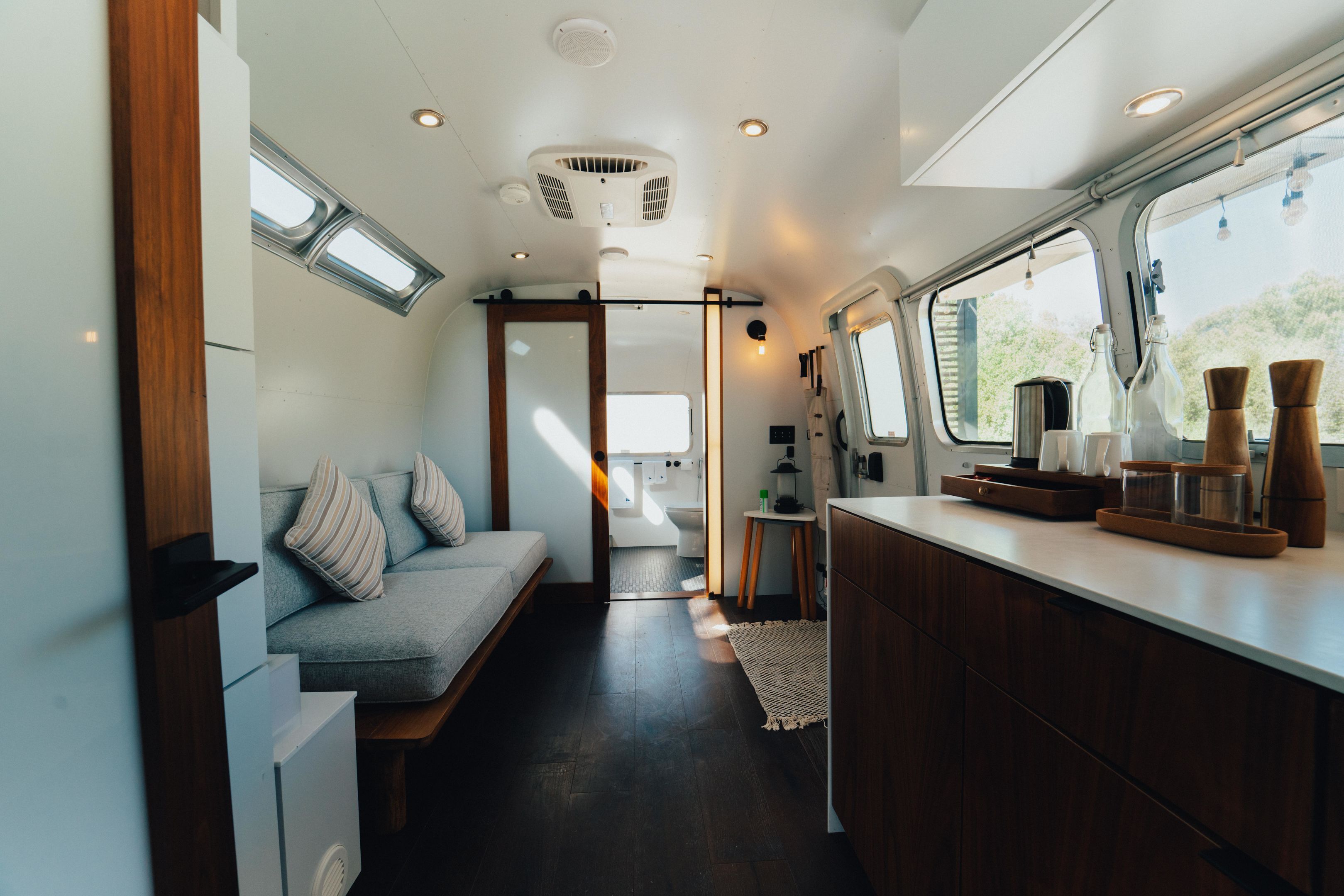 A compact, modern RV interior with a kitchenette, a seating area with cushions, and a window providing natural light.