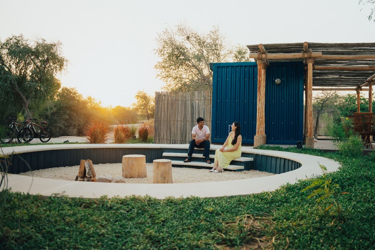 A circular outdoor seating area with wooden stools, a covered wooden structure, and a blue building in the background.