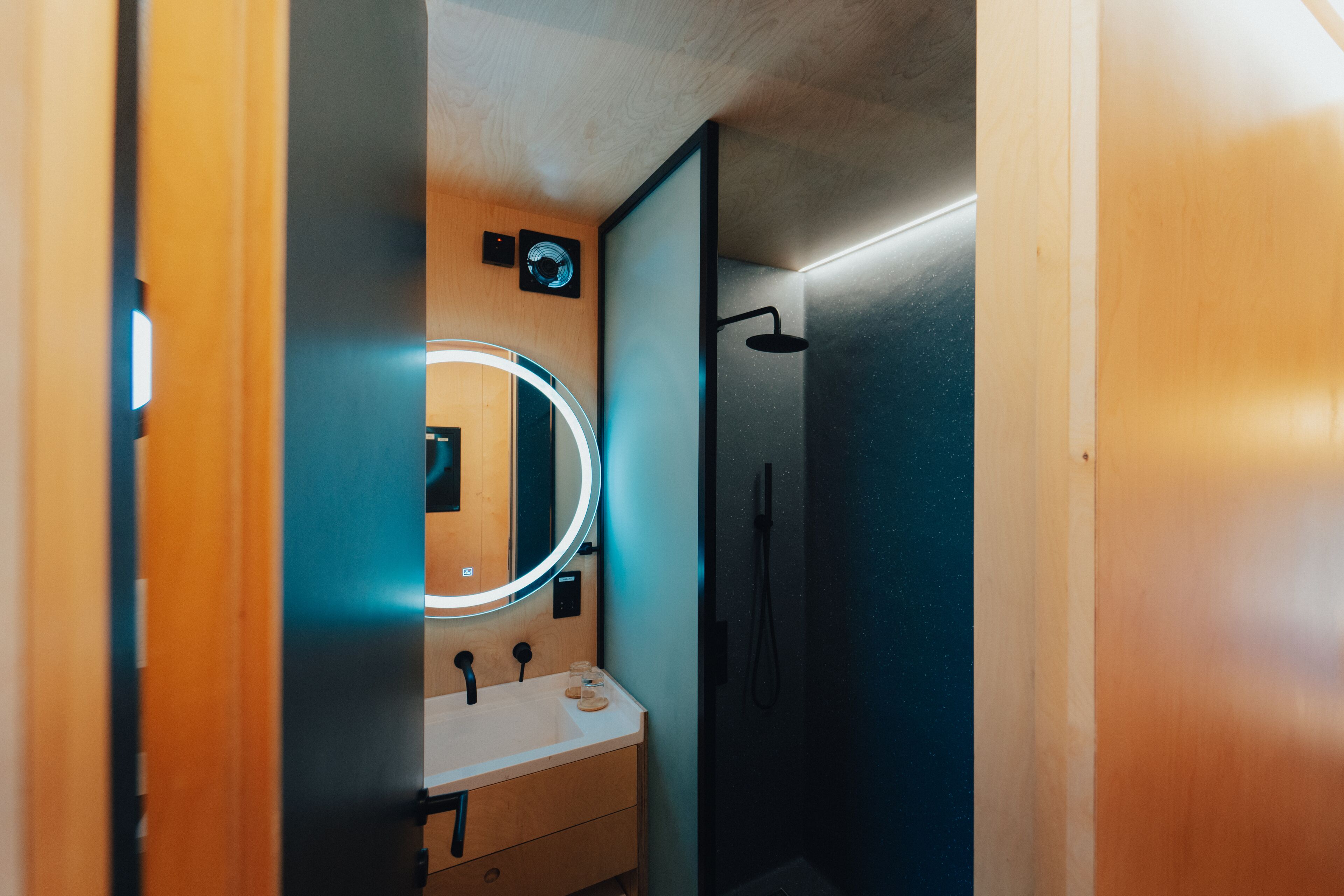 Une salle de bain avec un meuble-lavabo en bois, un miroir rond avec un éclairage intégré et un espace douche.