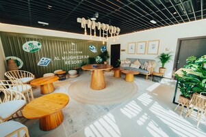 Um espaço de lounge moderno e bem iluminado, com mesas de madeira redondas e assentos confortáveis, apresentando uma parede verde com o logotipo e nome do resort.