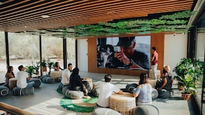 A group of people seated on circular stools in a modern, open-plan space with a large screen displaying a video. -  Bab Al Nojoum Al Mugheirah (Al Mirfa)