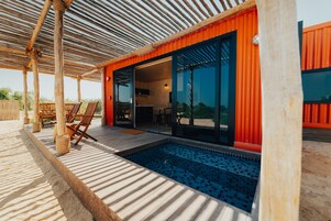 Uma área de piscina moderna ao ar livre, com deck de madeira e espreguiçadeiras, conectada a um edifício com grandes portas de vidro e fachada vermelha.