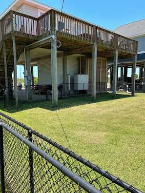 A covered outdoor area with a wooden deck, a fenced-in grassy lawn, and a building with a white exterior.