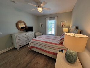 A bedroom with a bed, a dresser, a lamp, a ceiling fan, and a window with curtains. - Indian Bayou Lake House 2 - Lakeside Oasis (Fort Myers Beach)