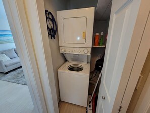A compact laundry room with a washing machine and dryer, a door leading to a living area, and a shelf with cleaning supplies. - Indian Bayou Lake House 1 - Lakeside Oasis (Fort Myers Beach)