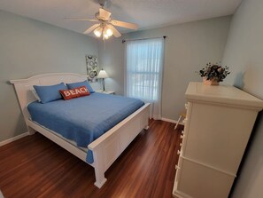 A bedroom with a bed, a ceiling fan, a nightstand, a lamp, a window with blinds, and a plant.