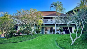 A two-story house with a balcony, surrounded by well-maintained lawns and tropical plants.