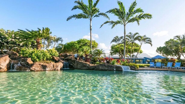 A clear pool with a waterfall, palm trees, and a lounge area with blue chairs.