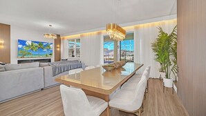A modern living room with a large sofa, a dining table with chairs, and a large TV screen displaying a tropical scene.