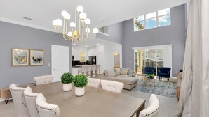 A spacious dining area with a large wooden table and white upholstered chairs, a chandelier, and a view into the kitchen and living room.