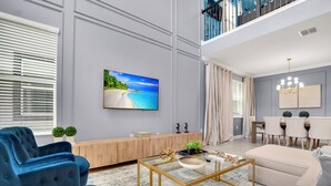 A modern living room with a large TV, blue sofas, a glass coffee table, and a dining area with a chandelier.