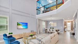 A spacious living room with a large sofa, a coffee table, and blue armchairs. A TV is mounted on the wall, and there is a staircase leading to an upper level.