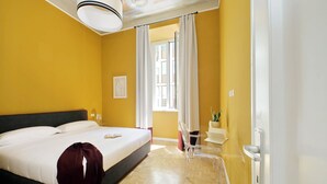A hotel room with a bed, a bedside table, a chair, a window with curtains, and a view of the outside. - Monti Apartments - M5 (Rome)