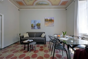 A living room with a sofa, a glass dining table, and chairs. There are two paintings on the wall and a plant on the table.