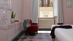 A bedroom with a bed, a red chair, a radiator, a window with curtains, and a view of a balcony with plants.