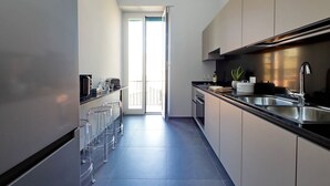 A modern kitchen with a stainless steel refrigerator, a black countertop, and a sink. There is a dining area with clear glass chairs and a white table.