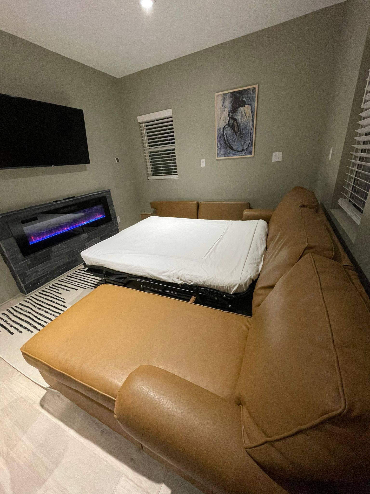 A modern living room with a brown leather sofa, a flat-screen TV mounted on the wall, a fireplace with a glass front, and a painting on the wall.