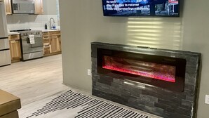 A modern living room with a flat-screen TV mounted on the wall, a stone fireplace, and a patterned rug. - Cozy Modern Home  (Lake Placid)