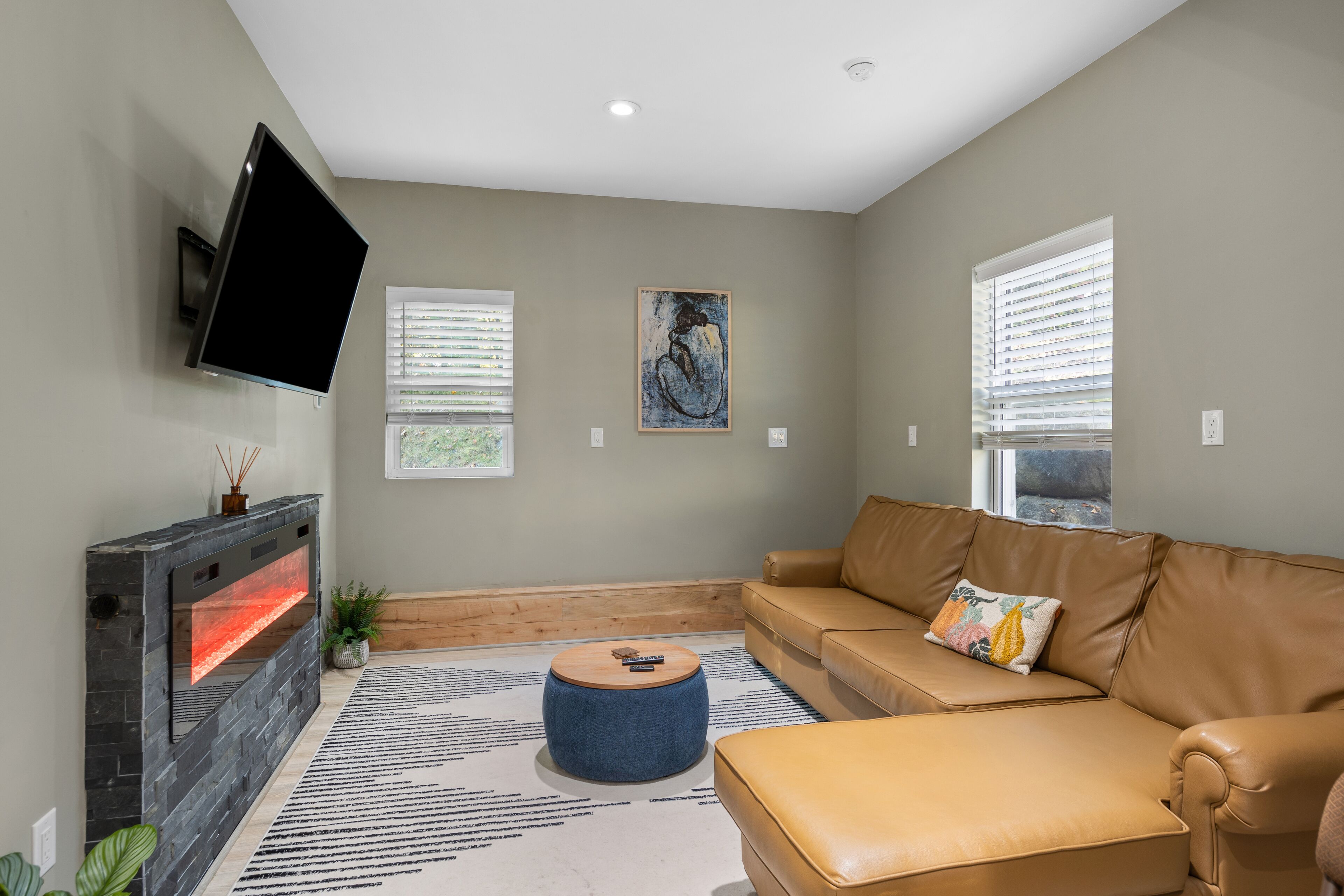 A living room with a stone fireplace, a leather sofa, a round ottoman, and a flat-screen TV mounted on the wall.