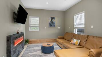 A living room with a stone fireplace, a leather sofa, a round ottoman, and a flat-screen TV mounted on the wall.