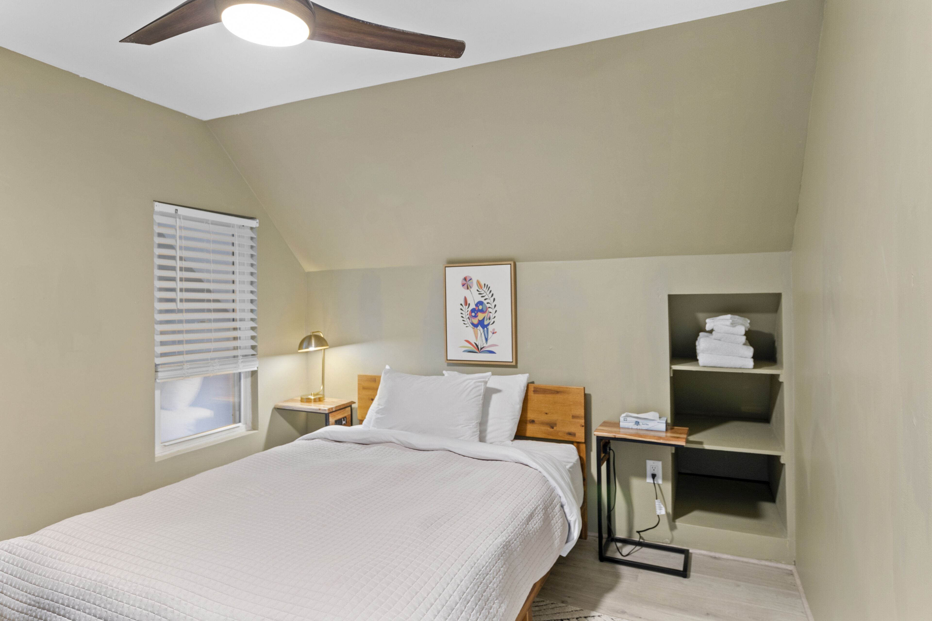 Une chambre à coucher comprenant un lit, une table de chevet, une étagère fixée au mur avec des serviettes, un ventilateur de plafond et une fenêtre avec des stores.