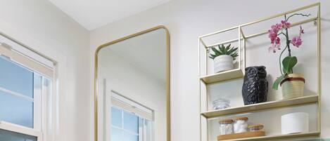 A bathroom with a white sink, wooden vanity, and a toilet. There is a mirror above the sink and a shelf with plants and a vase.